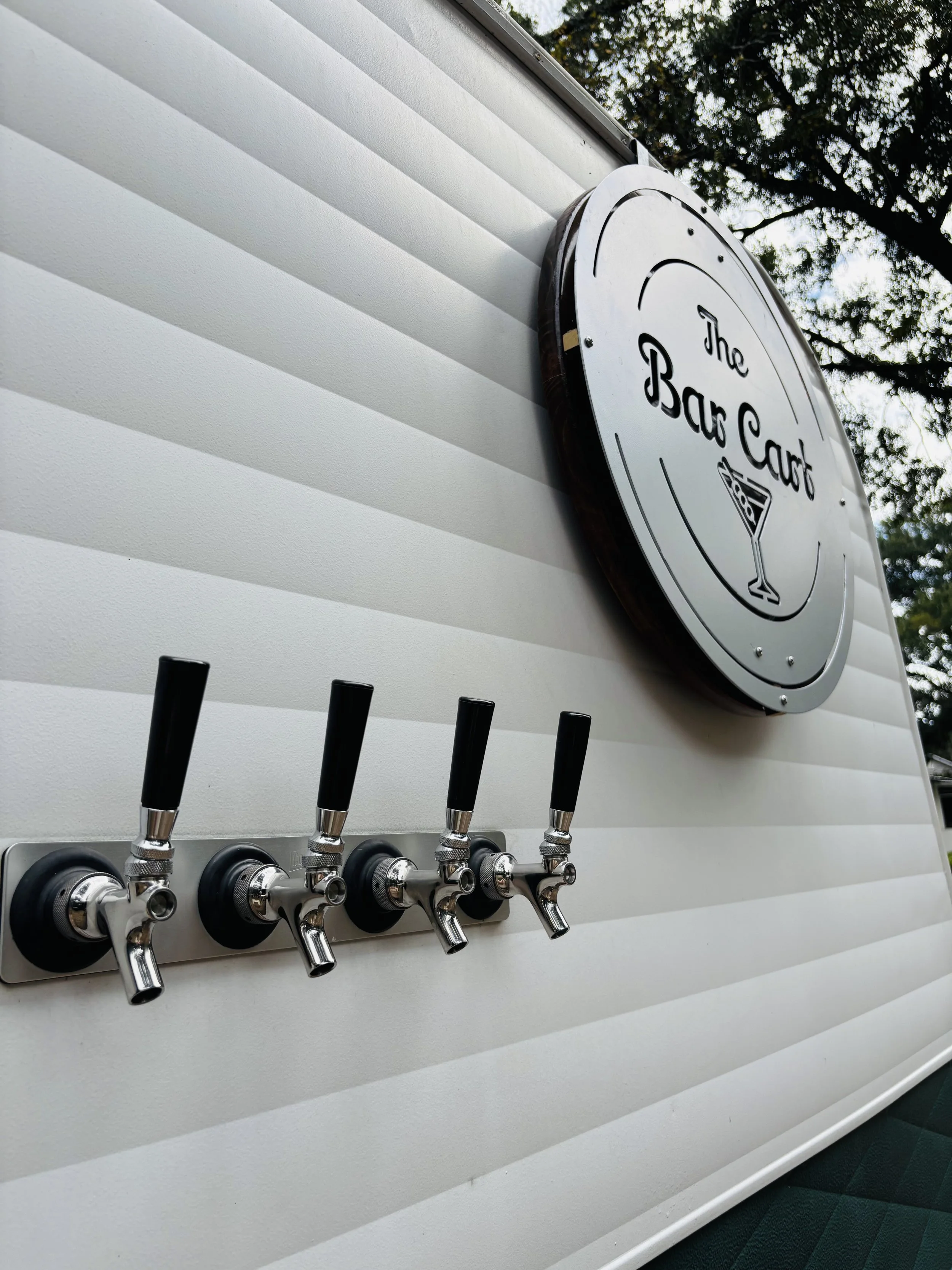 A close-up of a bar cart with five beer taps mounted on a white wall and a sign reading 'The Bar Cart' with a martini glass illustration.