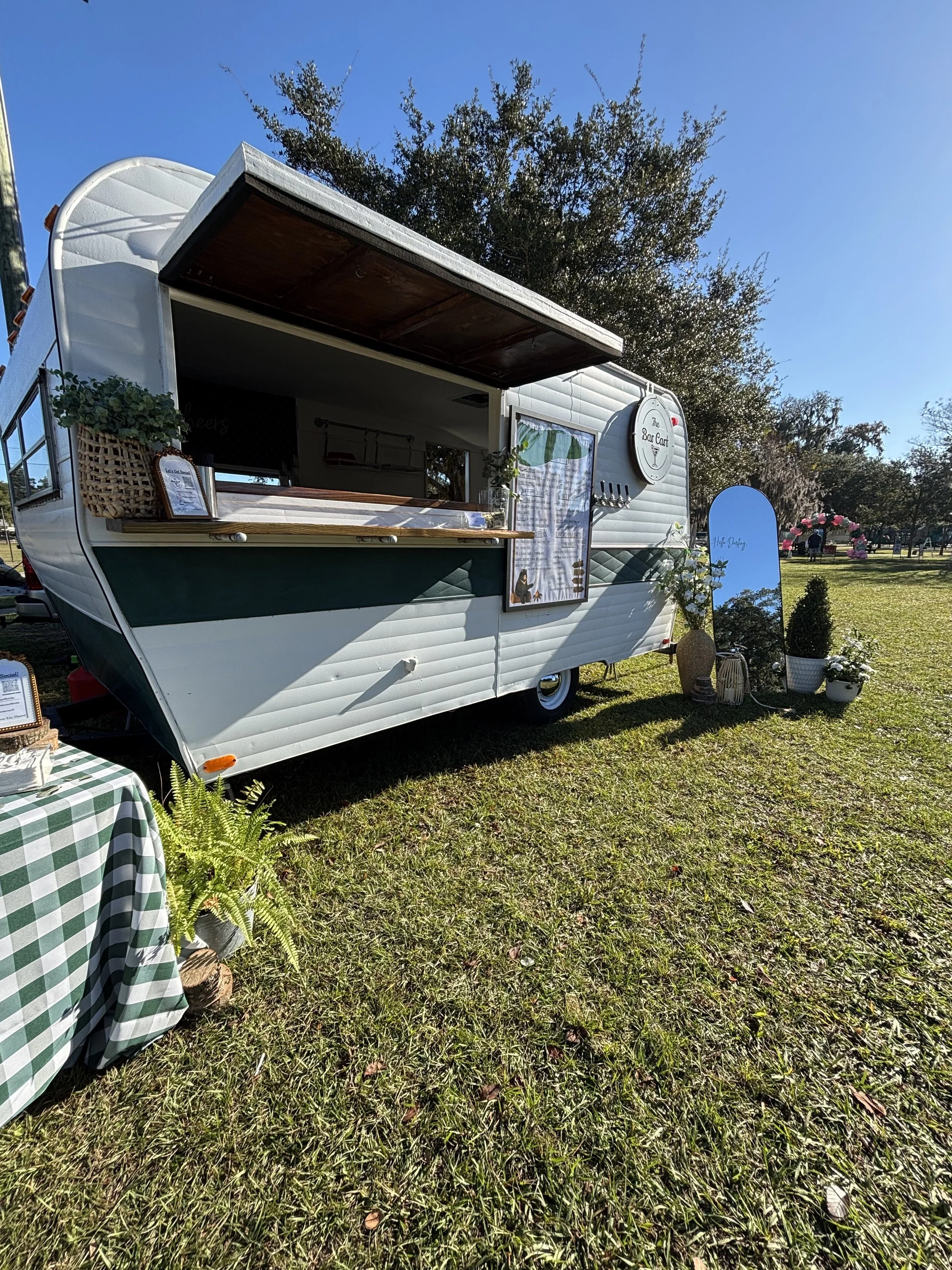 A white vintage trailer set up as a mobile bar with a serving window, outdoor decorations, and potted plants on the grass in a park.