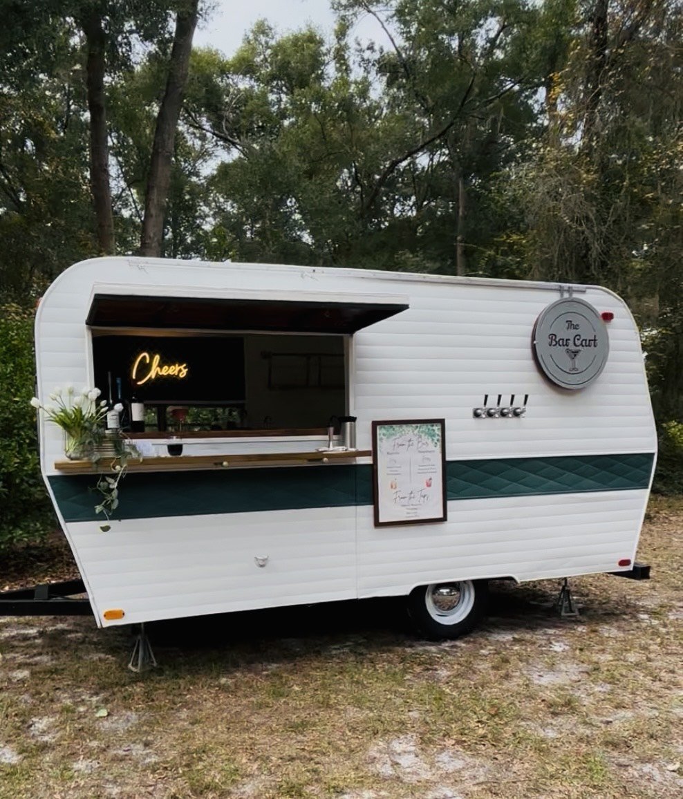 A small white mobile bar trailer with a green stripe, set up outdoors among trees. It has a serving window with a wooden counter displaying a flower vase and drinks, and a sign reading 'Cheers'. The trailer is branded as 'The Bar Cart' with a circula