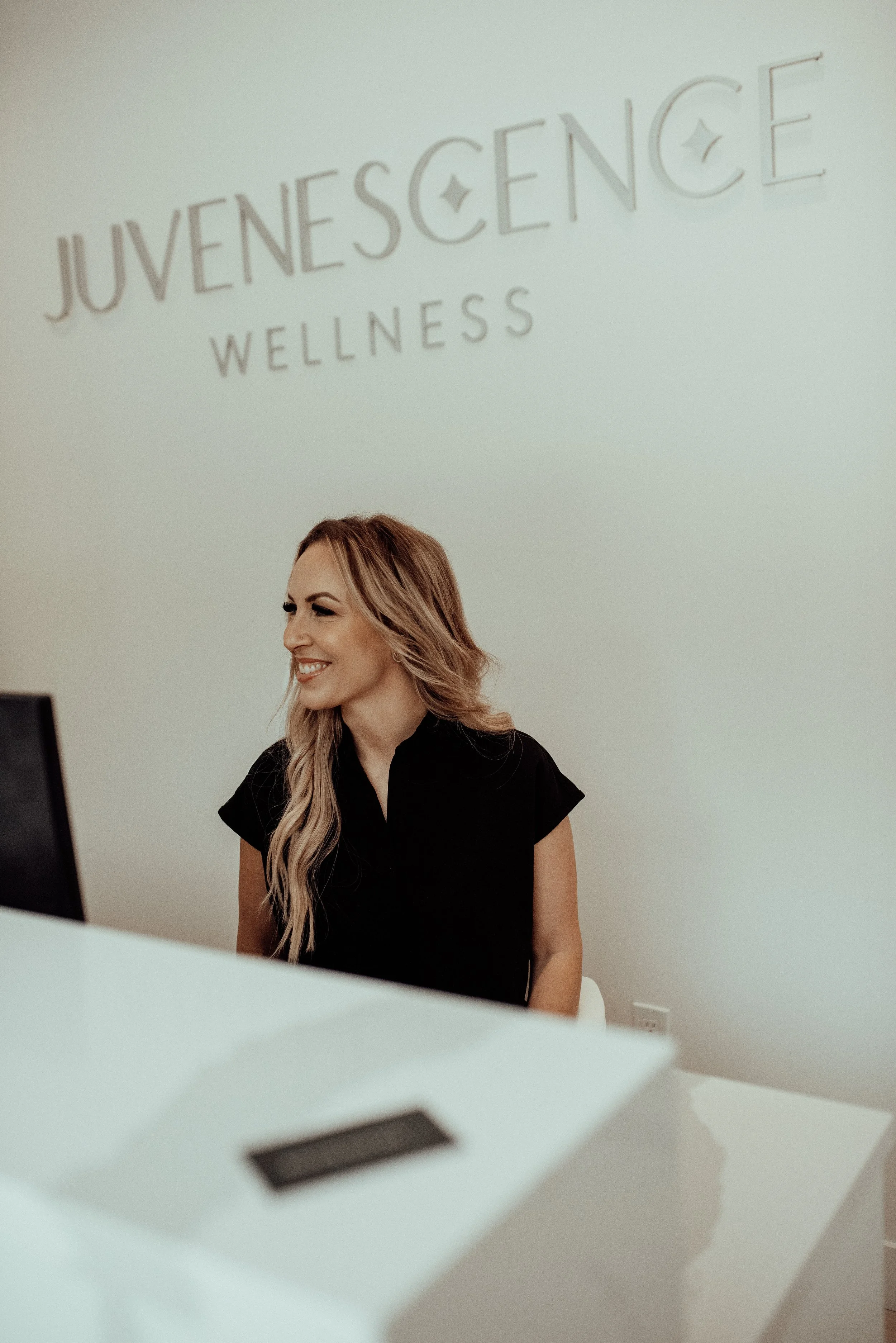 A woman sitting at a reception desk smiling, with a sign on the wall behind her that reads "JUVENESCENCE WELLNESS".