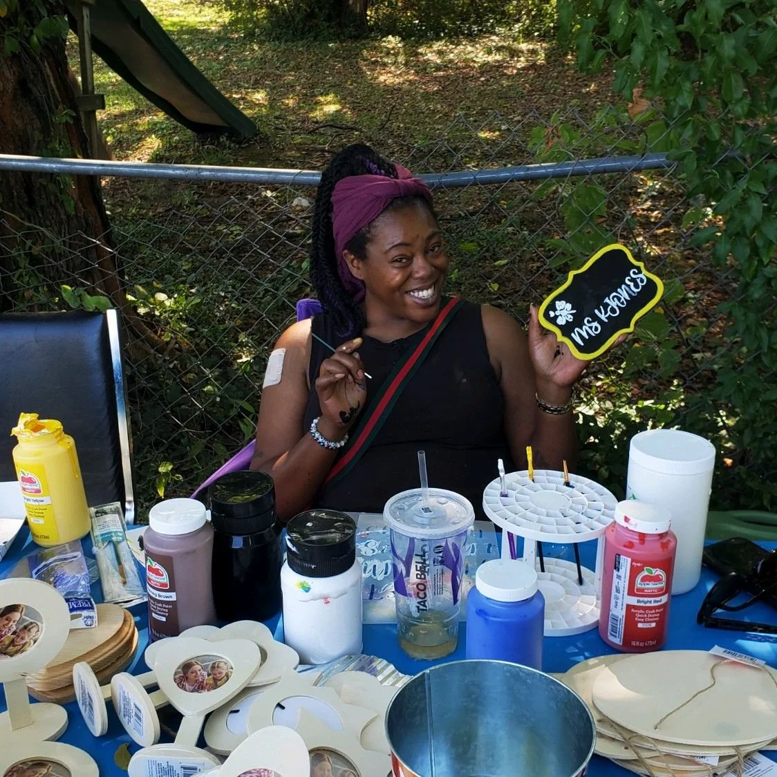 A woman sitting at a table outdoors, smiling and holding a small chalkboard sign that says 'Ms. Kianse.' The table has supplies like bottles of paint, cups, and wooden heart-shaped photo frames.