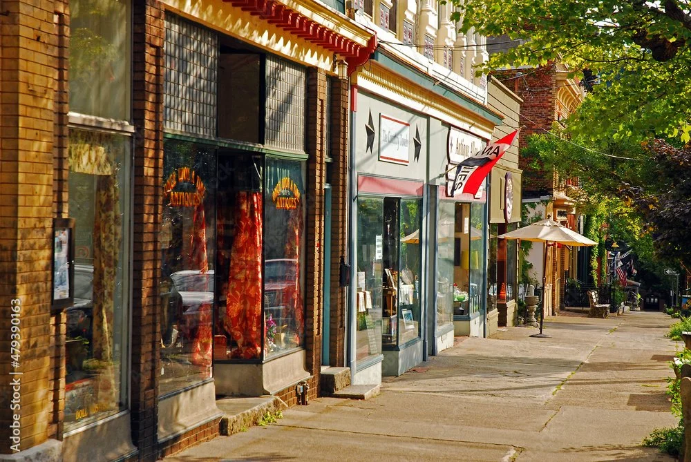 A sidewalk scene in a small town with a row of storefronts, including antique shops, under leafy trees with a few outdoor seating areas and umbrellas.