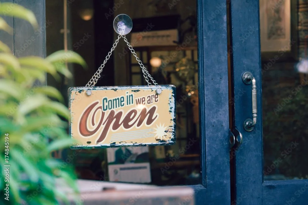 A vintage-style sign hanging on a glass door reads 'Come in we are Open'. The door has a silver handle and the interior of the store is slightly visible through the glass.