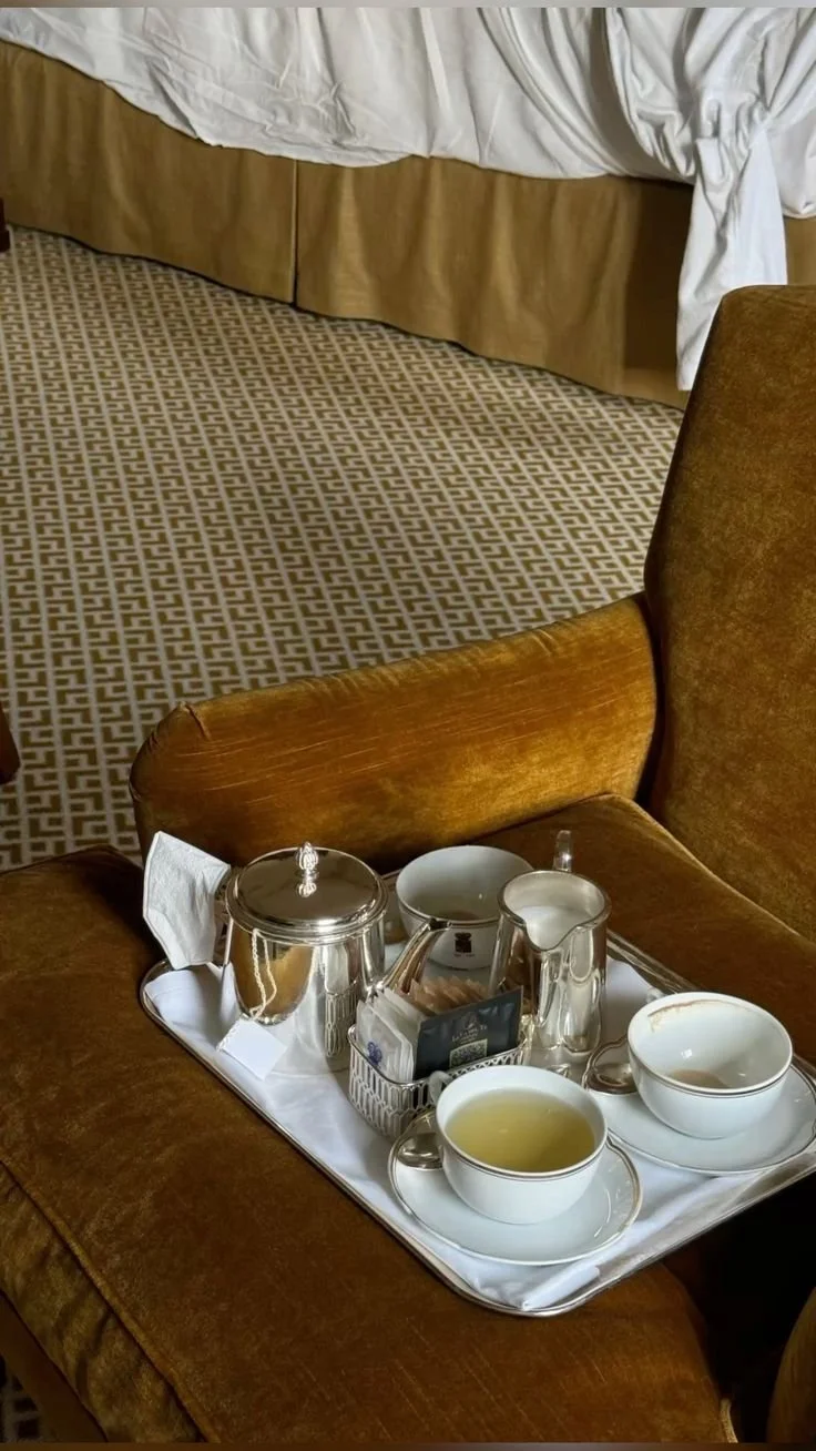 Empty tray with a tea set, including a teapot, cups, creamer, and sugar container, placed on a brown velvet armchair in a hotel room.