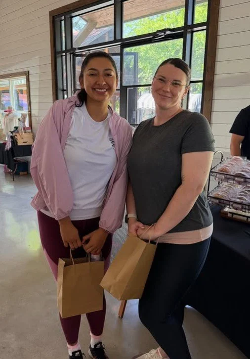 Two women standing inside a room, smiling, holding brown paper bags, near a window with sunlight outside, in a casual setting with tables and items in the background.