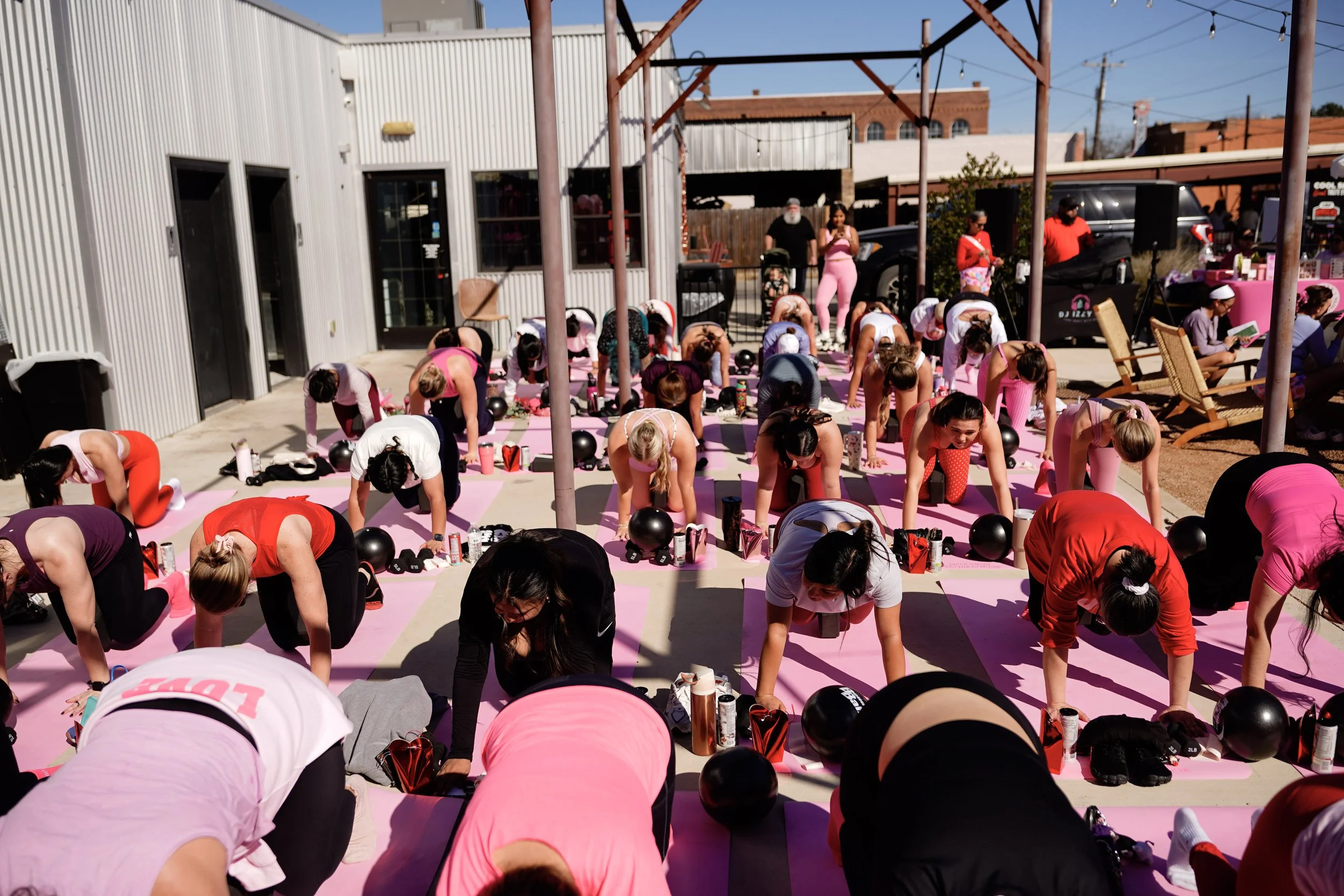 A group of women participating in an outdoor yoga class on pink mats, with some using water bottles and black balloons, in front of a building with rustic brick and metal siding, under a clear blue sky.