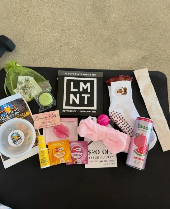 Collection of personal care and gift items on a black surface, including a green bath bomb, a black and white LMNT electrolyte drink mix, a white sock with a small embroidered design, a pink plush headband, a pink yo-yo, a can of Strawberry-flavored 
