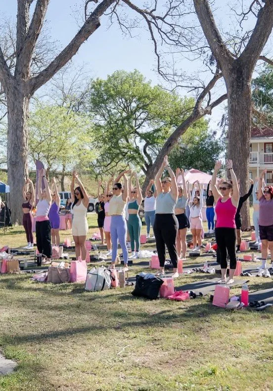 Group of women participating in outdoor yoga session under trees, stretching with arms raised, yoga mats and bags on the grass.