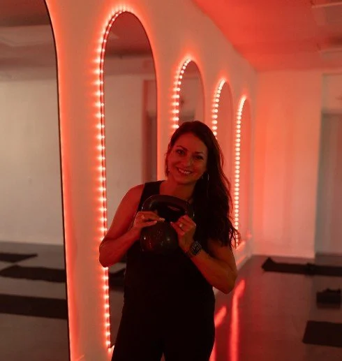 Woman with long dark hair taking a selfie in front of a brightly lit red archway with string lights inside. She is smiling and wearing a black sleeveless top in a dimly lit indoor space with a black and white floor.