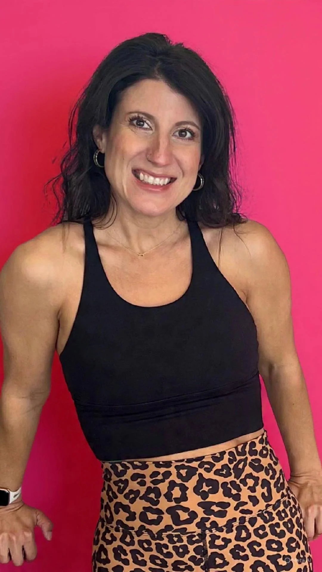 A woman with black hair, wearing a black sleeveless top and leopard-print pants, standing in front of a pink background.