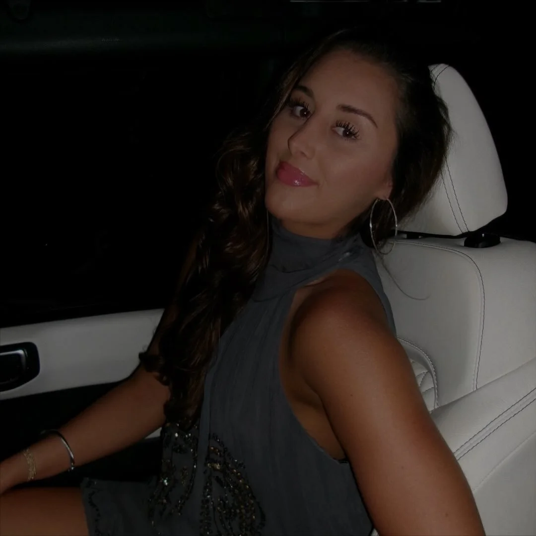 A woman with long, curly hair sitting in the backseat of a car at night, wearing hoop earrings and a sleeveless top.