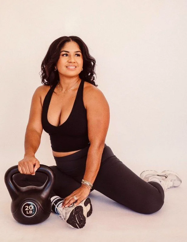 A woman in black workout clothing sitting on the floor, holding a 20-pound kettlebell, with a white background.