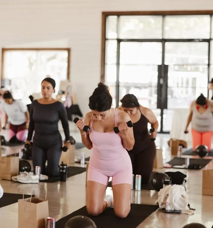 Women in a fitness class doing exercises with weights on kneeling mats in a bright gym with large windows.