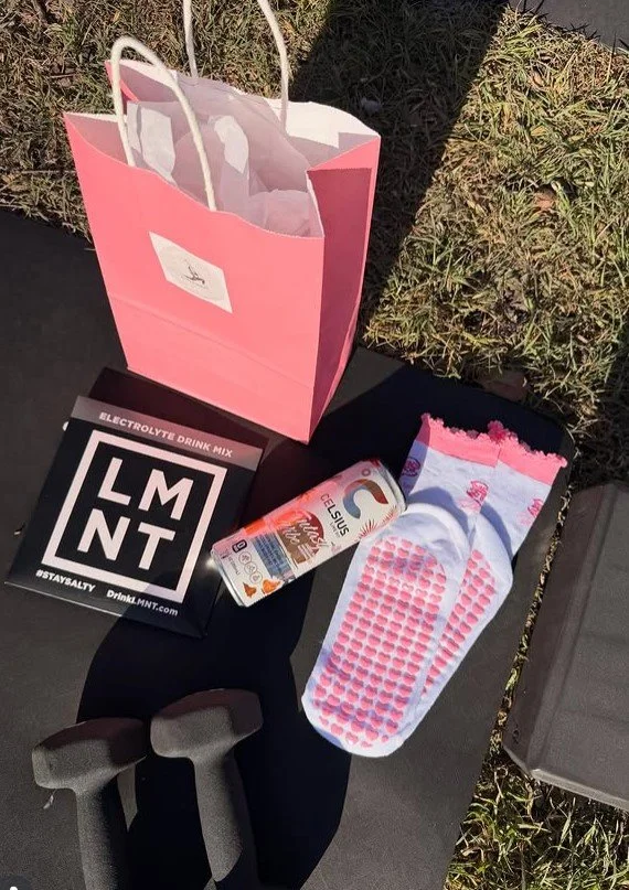 A pink paper gift bag, an electrolyte drink mix box, a can of coconut water, a pink and white athletic sock, and a black pair of sunglasses on a black surface outside next to grass.