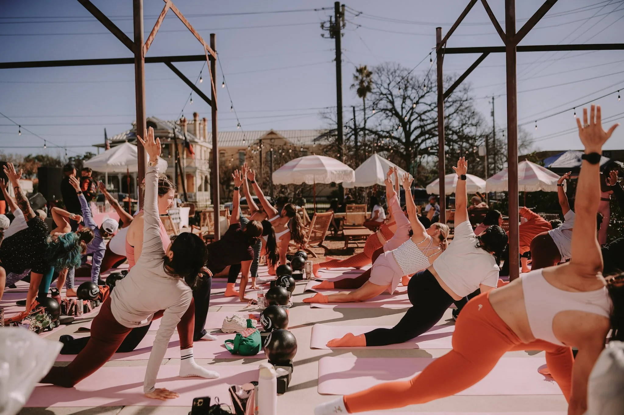 A group of people participating in a yoga class outdoors, doing stretches on yoga mats, with some raising their arms and others in various poses, under string lights and umbrellas on a sunny day.