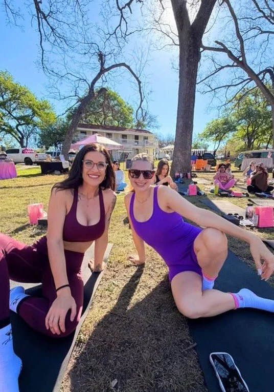 Two women in athletic wear doing yoga outdoors on mats in a park, smiling at the camera, with trees and other people practicing yoga in the background.