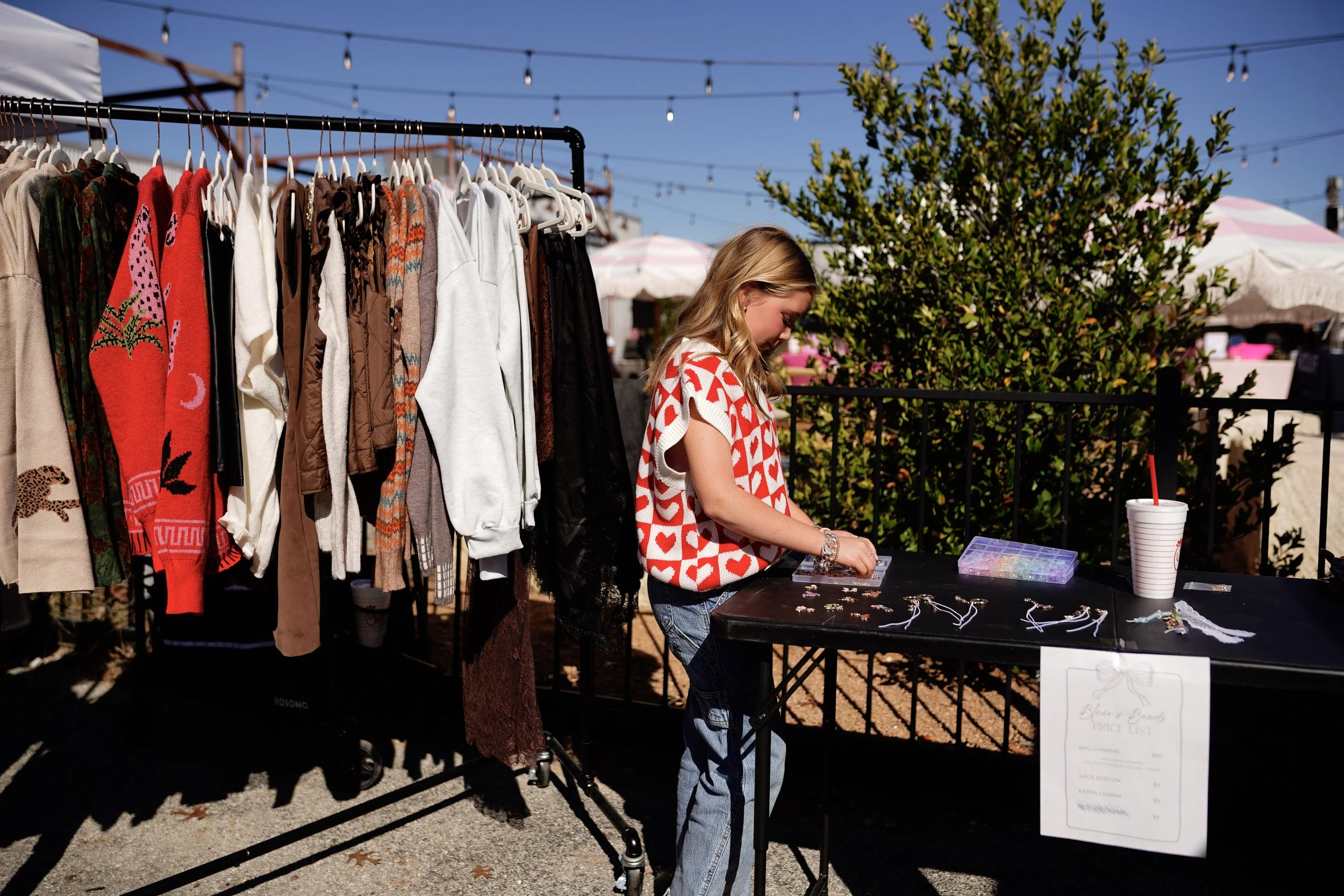 A young girl with blonde hair browsing jewelry on a black table at an outdoor market. Behind her, there are hanging sweaters and jackets on a clothing rack, and some pink and white umbrellas in the background with string lights above.