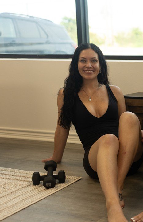 Woman with long dark hair sitting on the floor next to a dumbbell, smiling indoors near a window.