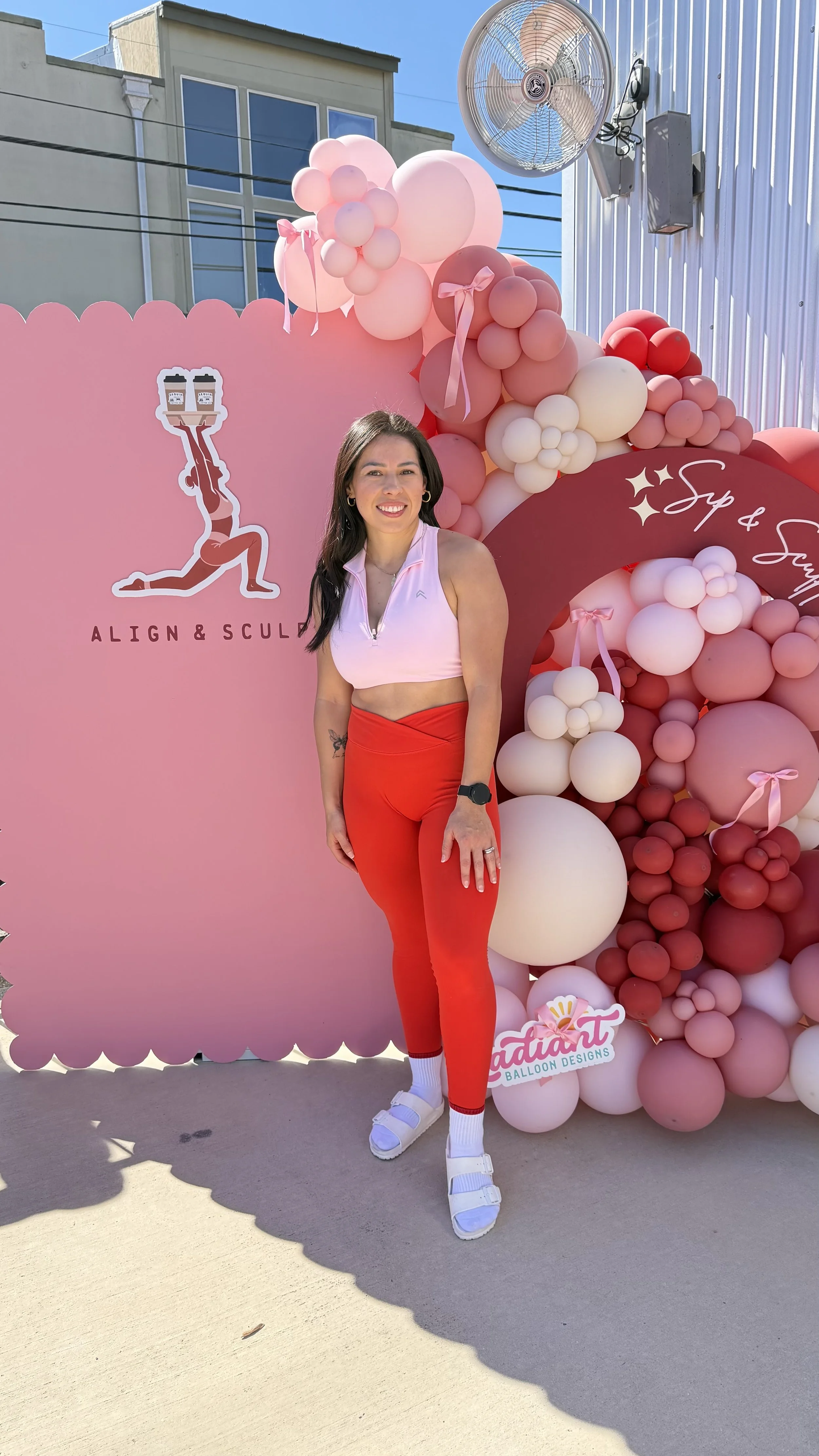 A smiling woman with long dark hair, wearing a white sleeveless top, red pants, white socks, and white sandals, standing in front of a pink balloon display with pink and white balloons. There is a pink sign with a woman holding two coffee cups and the text 'Align & Sculpt,' and a logo for 'Radiant Balloon Designs.' A large wall fan is mounted on the building behind her, and the setting appears to be outdoors with a clear blue sky.