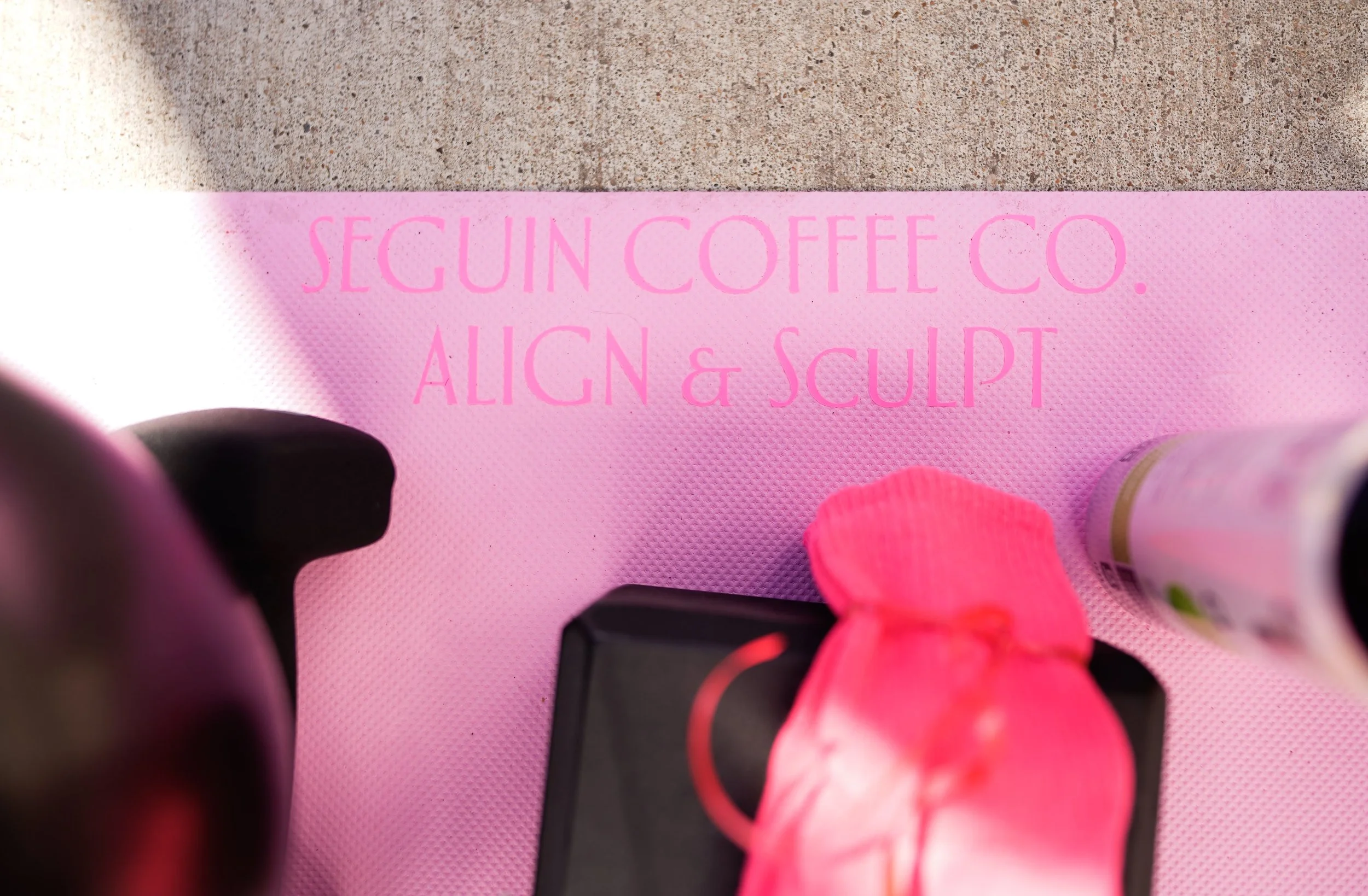 Pink mat with the text 'Secutr Coffee Co. Align & Sculpt' printed in pink. Part of a person's leg in a bright pink sock, black object, and a lotion bottle are visible.