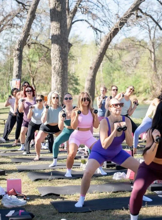 Group of women participating in outdoor fitness class, holding small weights, on yoga mats in a park with trees and a lake in the background.