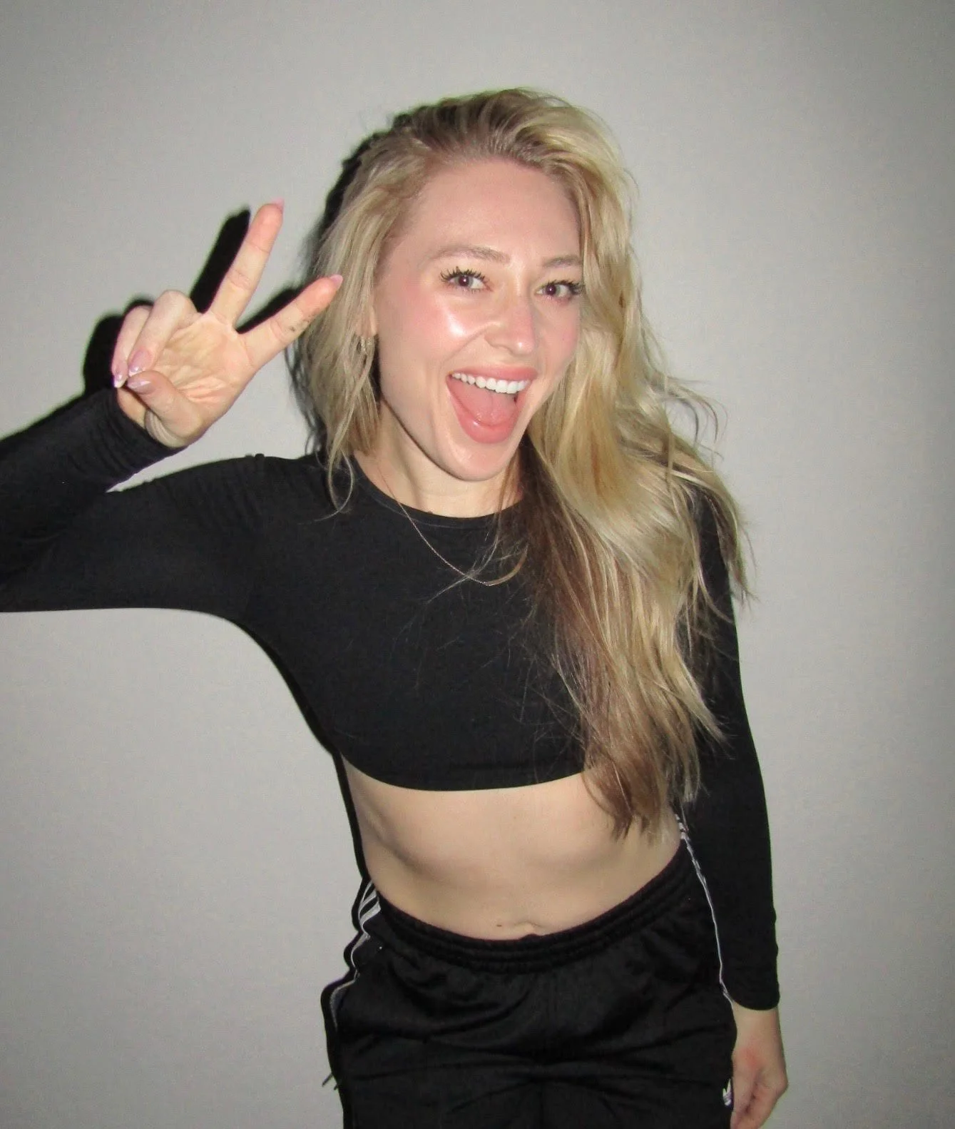 A woman with long blonde hair, wearing a black crop top and shorts, making a peace sign and smiling, standing against a plain wall.