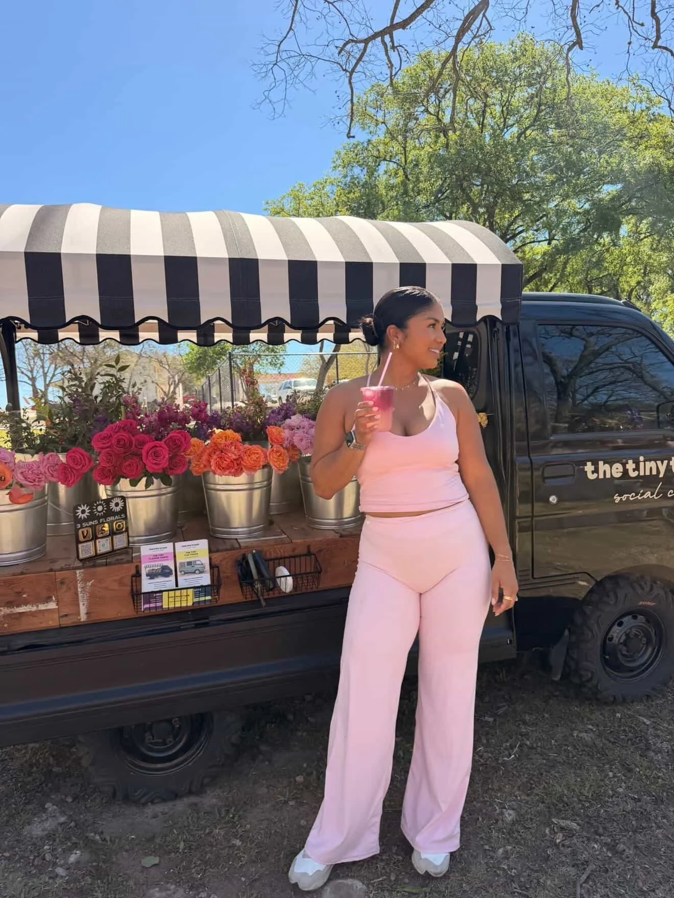 A woman in pink athletic wear holding a pink drink and standing next to a flower cart with pink, purple, and orange flowers on a sunny day in a park.