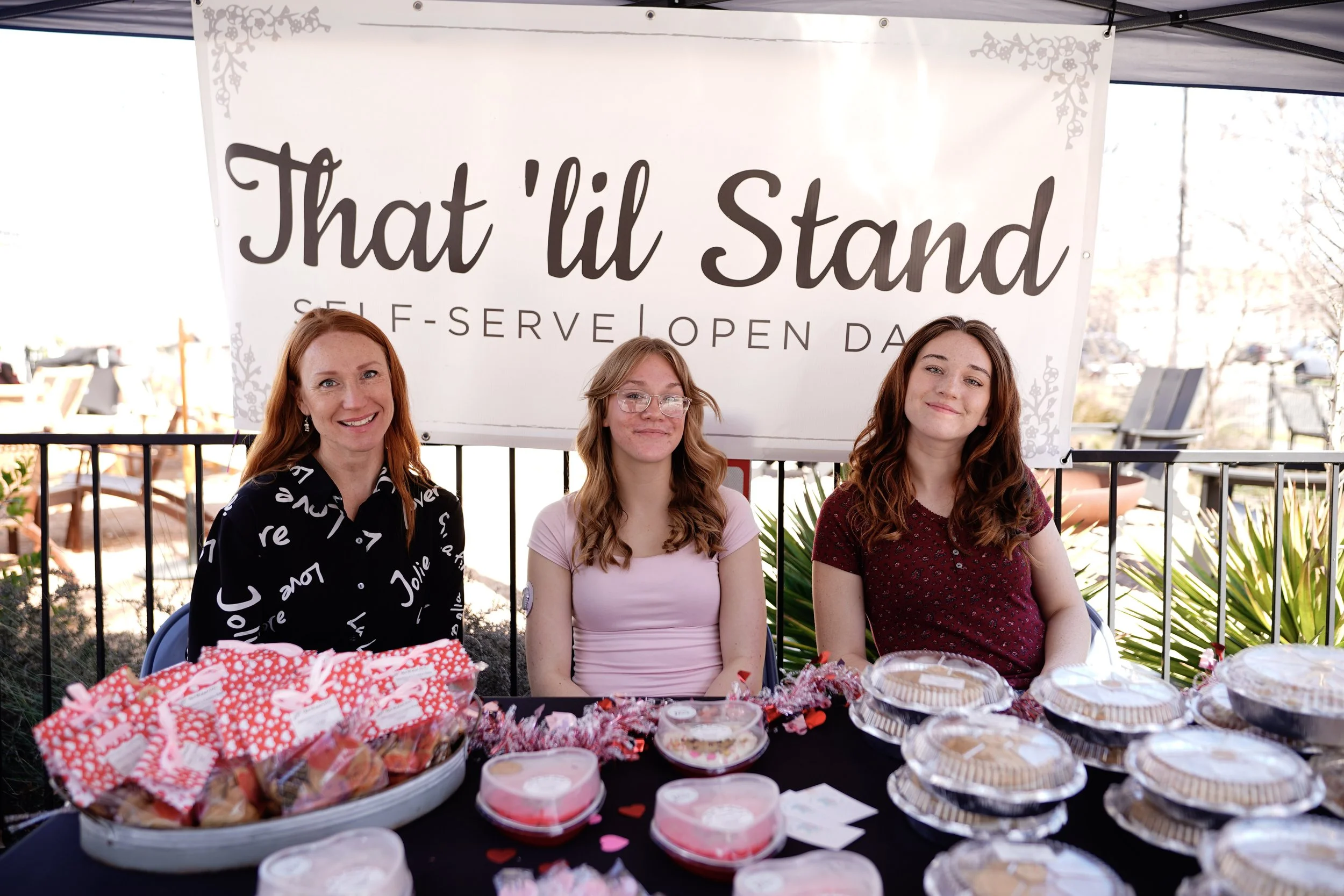 Three women sitting behind a table filled with baked goods, with a large sign behind them that reads 'That’d U Stand' and 'Self-Serve Open Daily'. The women are smiling and look happy, and the table is decorated with pink heart-shaped items, likely for Valentine's Day.