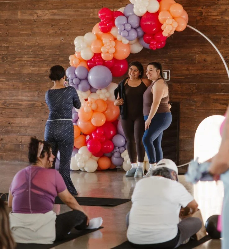 Three women stand in front of a large balloon arrangement of purple, pink, red, peach, and white balloons, taking a photo together at an indoor event, with some people sitting on the floor nearby.
