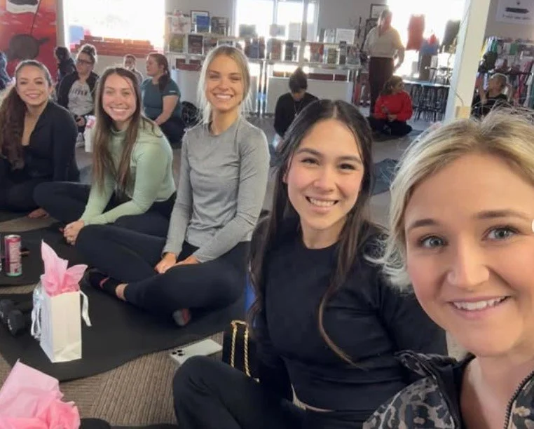 Group of five smiling women sitting on a black mat at a store or mall, with store shelves and other shoppers in the background.