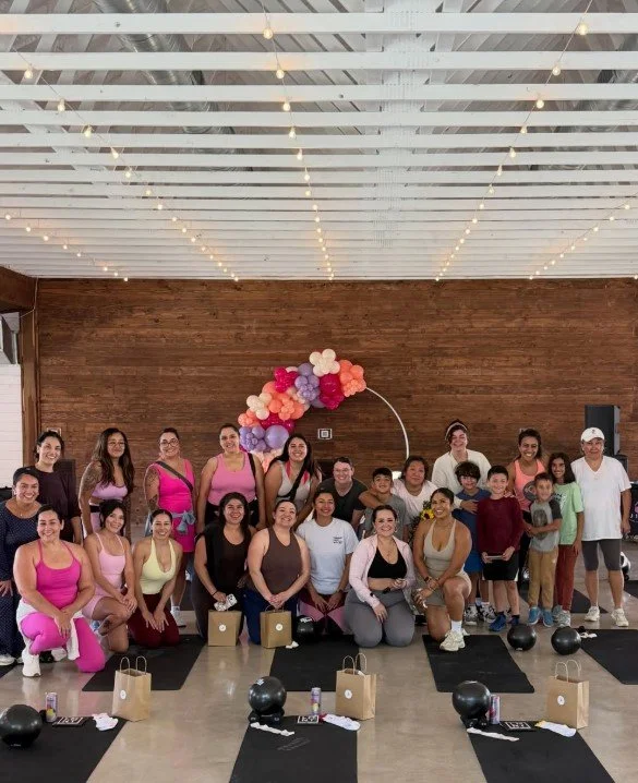 Group of people in a fitness class posing for a photo in a studio with wooden walls and string lights, with balloons forming an arch in the background.