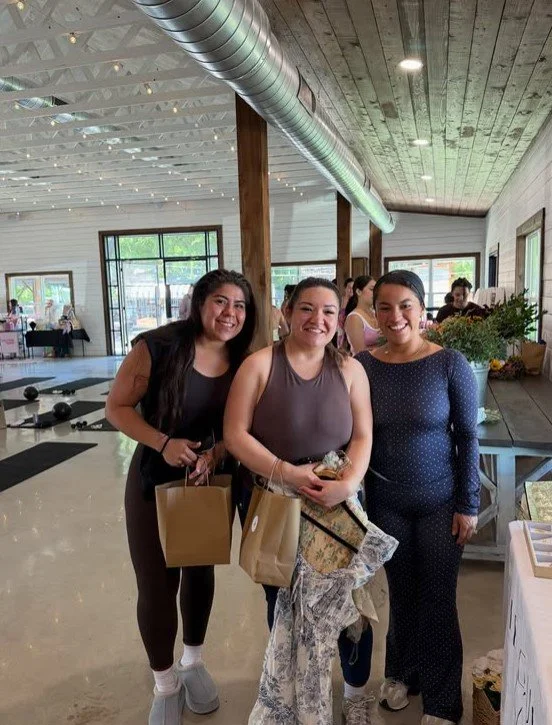 Three women smiling and holding gift bags inside a modern, well-lit building with wooden accents and large windows.