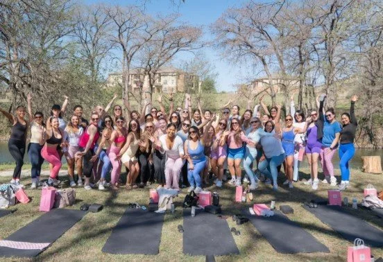 Group of women standing outdoors in a park, holding their hands in the air after a workout class, with yoga mats and bags on the ground.