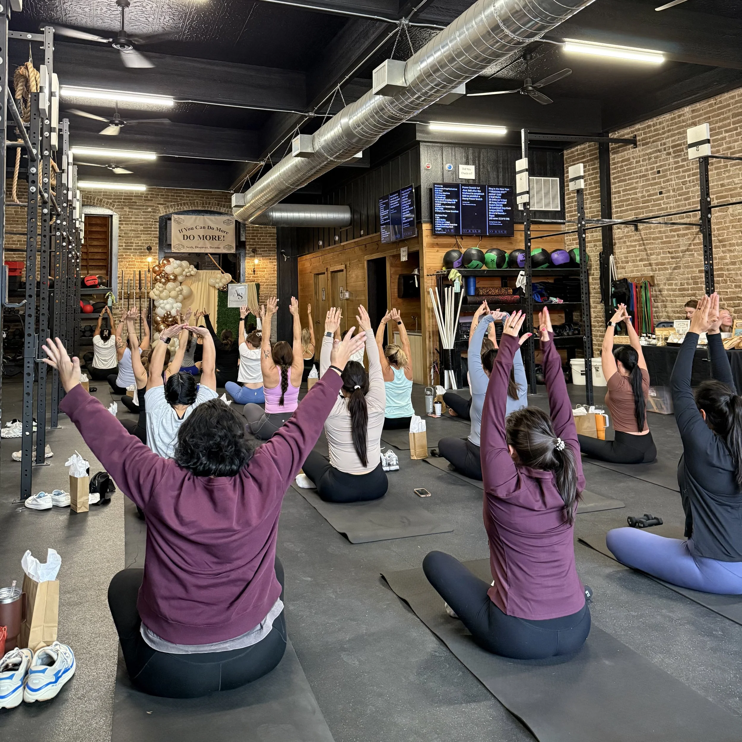 Interior of a fitness studio with people participating in a yoga or meditation class, sitting on black mats with arms raised, surrounded by workout equipment, brick walls, and digital screens displaying class information.