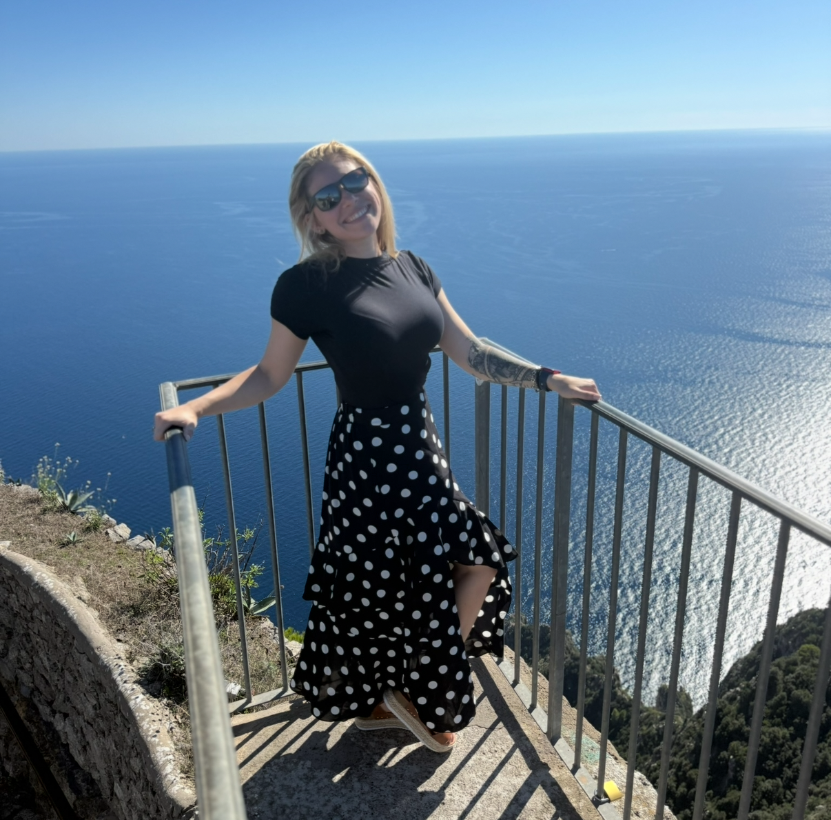 A woman with sunglasses and a tattoo on her arm posing at a seaside lookout with a metal railing, wearing a black top and a black and white polka dot skirt, smiling with the ocean in the background.