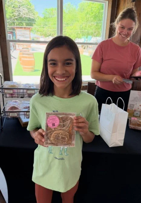 A young girl with shoulder-length dark hair smiling and holding a baked good in clear plastic wrap. Standing indoors near a table with baked goods and a woman in a pink t-shirt behind her, with a large window showing a backyard outside.