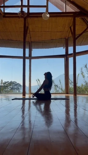 A woman practicing yoga in a spacious room with large floor-to-ceiling windows showing an outdoor landscape and mountains.