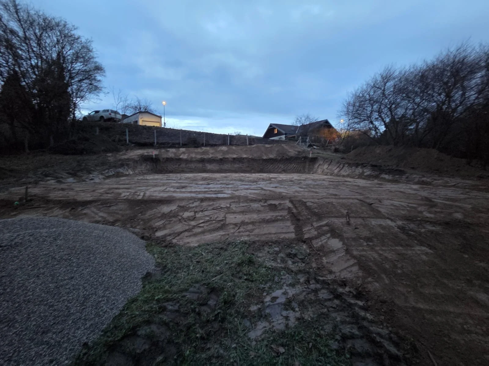 Baugrundstück unter hellem Himmel mit sichtbaren Erdboden, Hügeln, Bäumen und Häuser im Hintergrund.