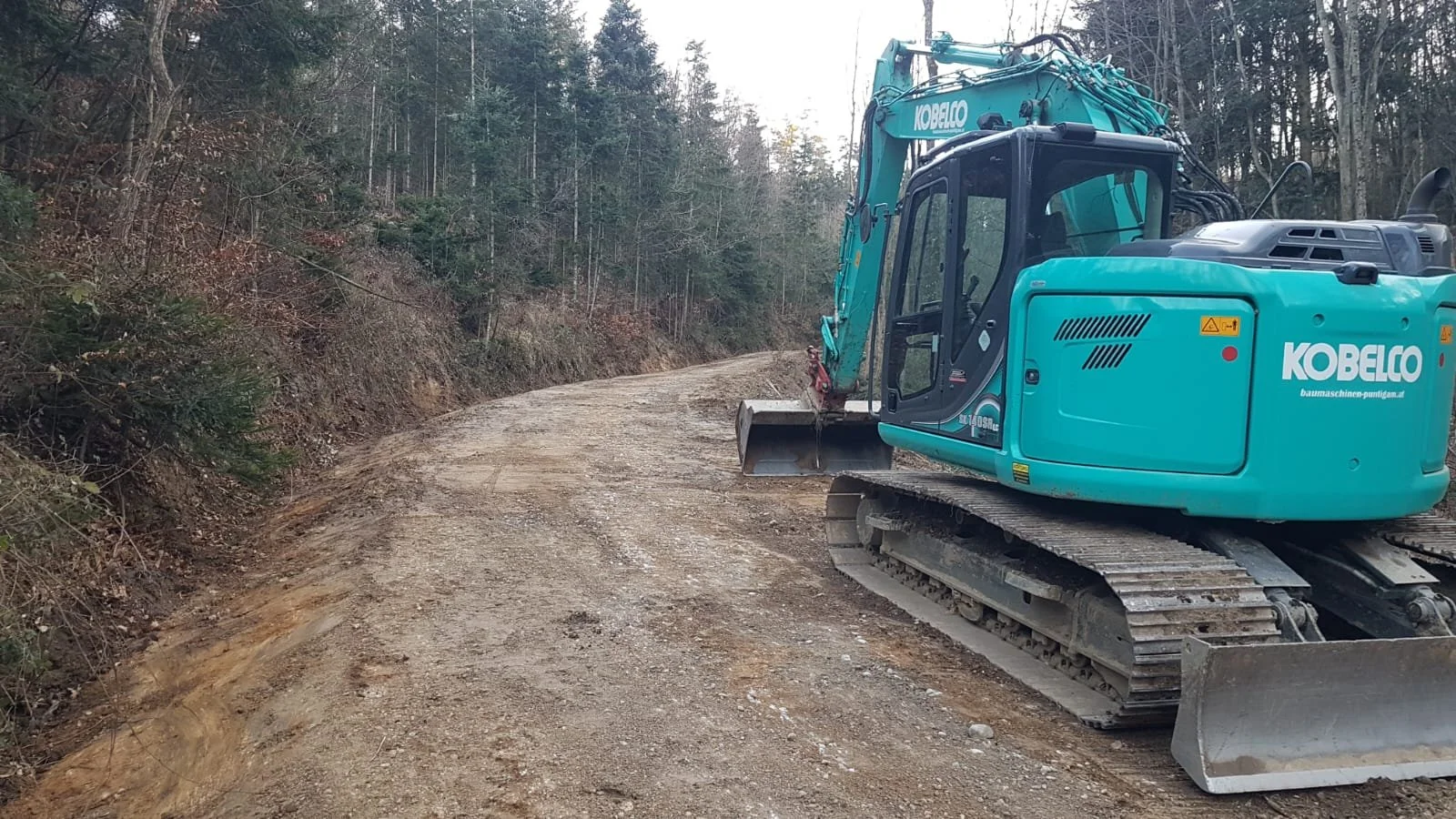 Ein blauer Bagger beim Bau eines unbefestigten Straßenabschnitts in einem Waldgebiet.