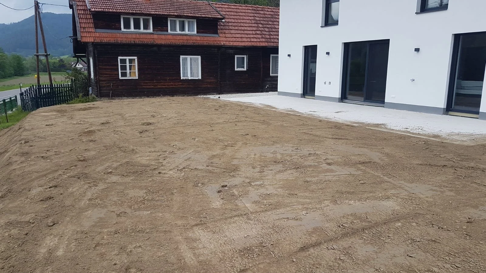 Ein umzäuntes Grundstück mit Erde, das für eine Gartenanlage vorbereitet wird, befindet sich neben modernen weißen und traditionellen Holzhäusern in einer ländlichen Gegend mit Bergen im Hintergrund.