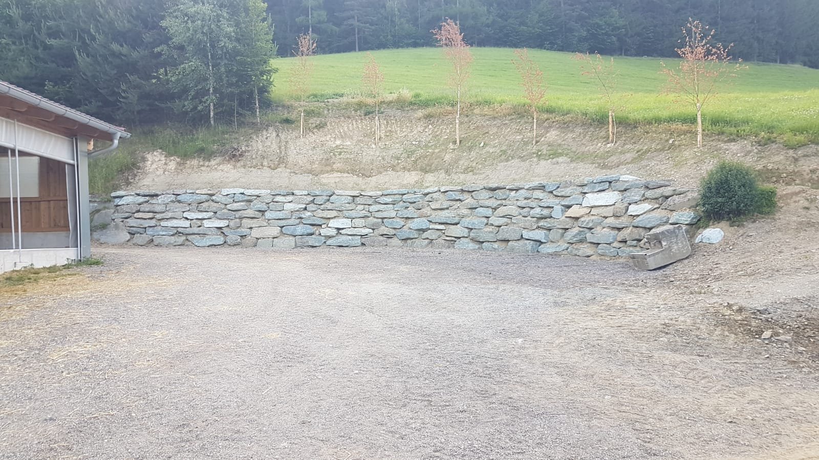Eine Terrassenmauer vor einem grünen Hügel mit Bäumen und kleineren landwirtschaftlichen Flächen.