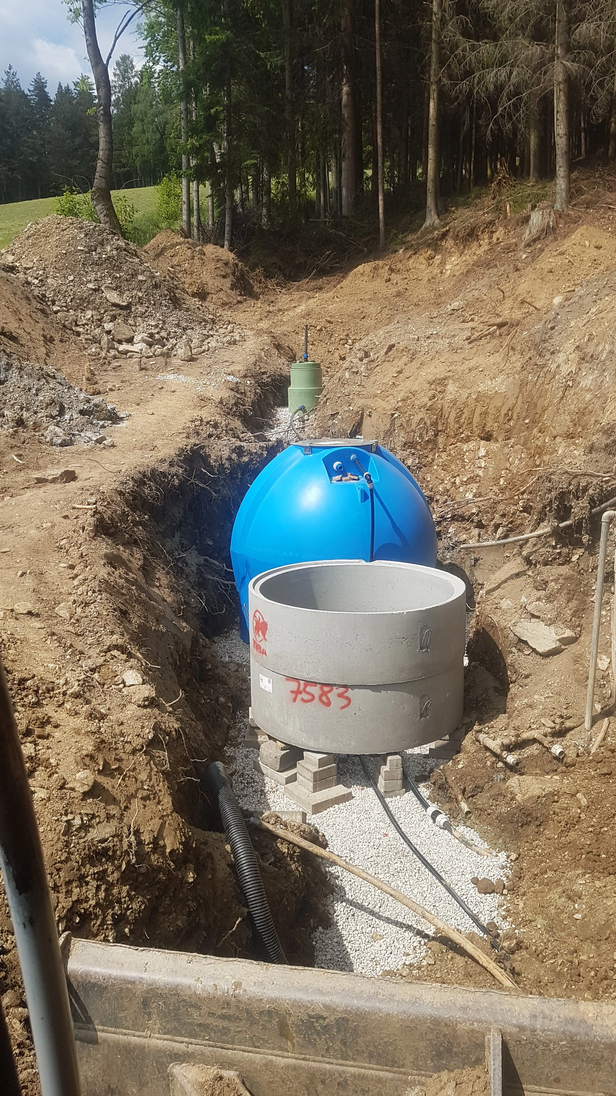 Bauarbeiten im Freien, Erdbau mit Rohren, Tank und Bauteilen in einer Grube, umgeben von Bäumen.