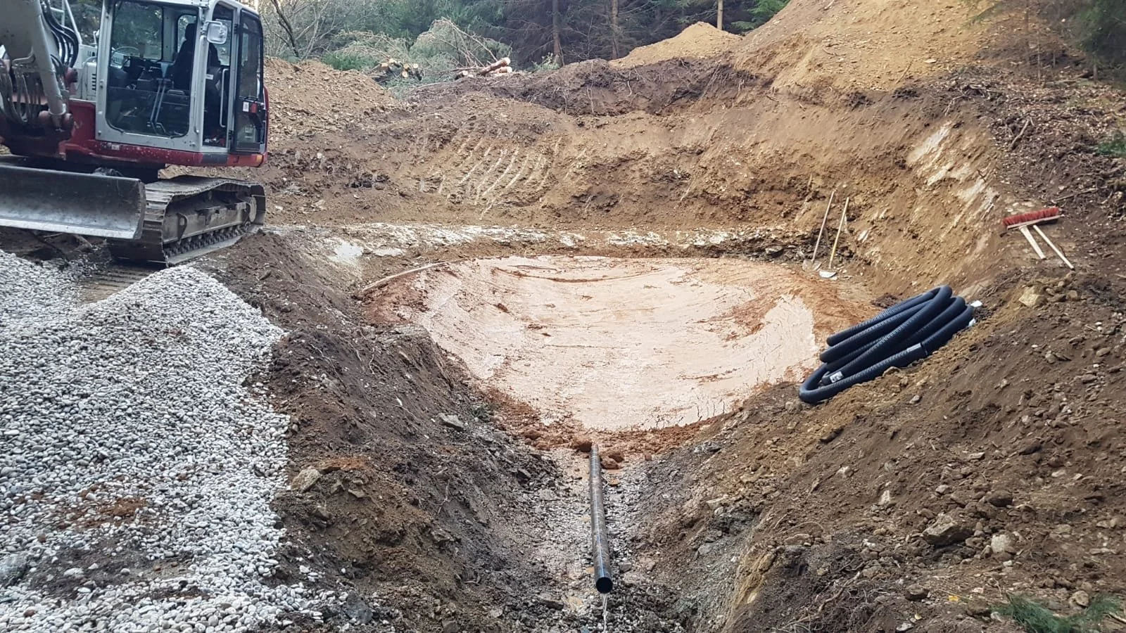 Ein Bauplatz mit Aushub, ein Bagger und Rohre vor einer Erdrückwand,bereit für den Bau eines Pools oder Teichs.