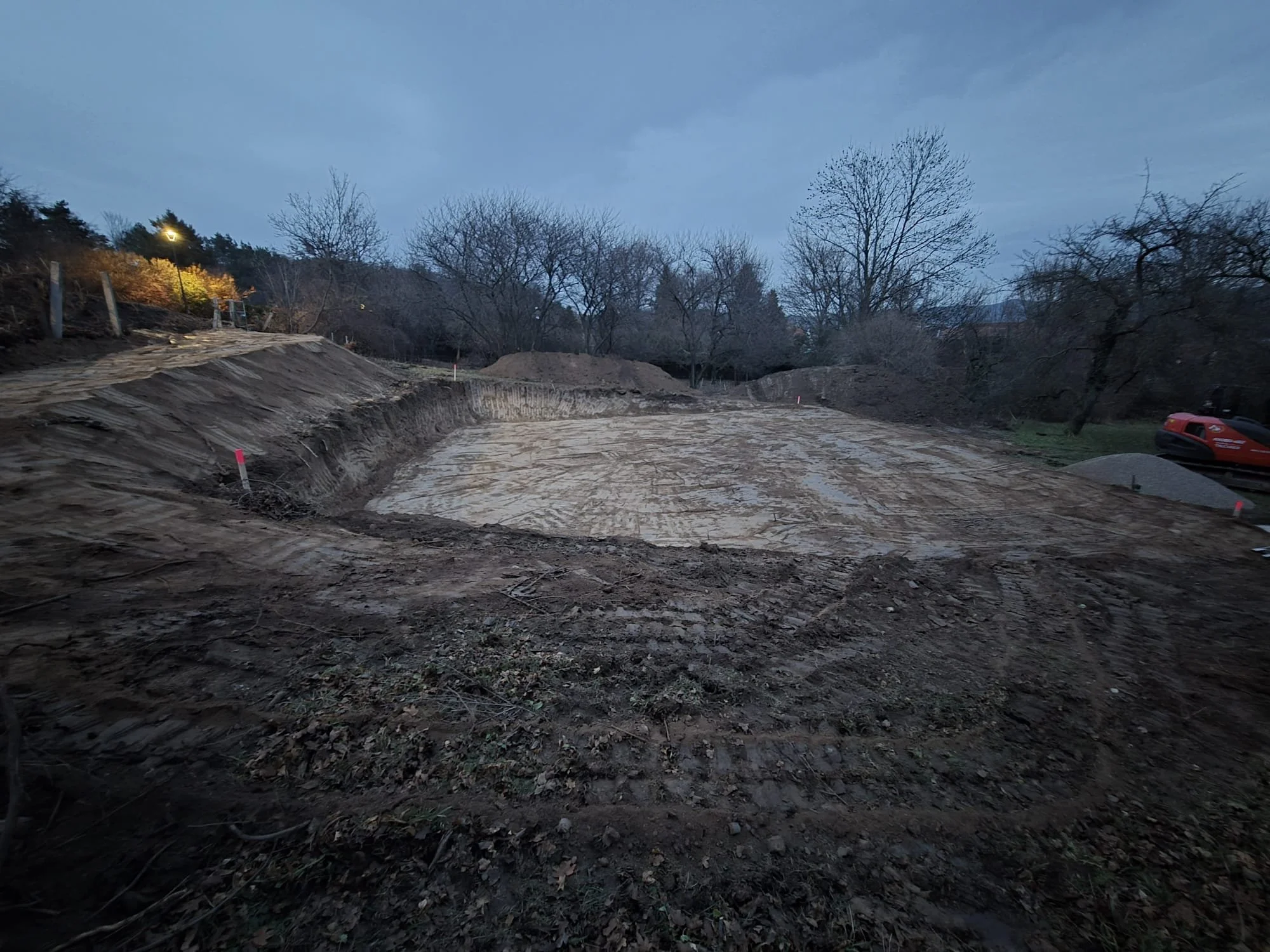 Baustelle mit ausgegrabtem Boden und Baugeräten, umgeben von Bäumen, während es draußen dunkel ist.