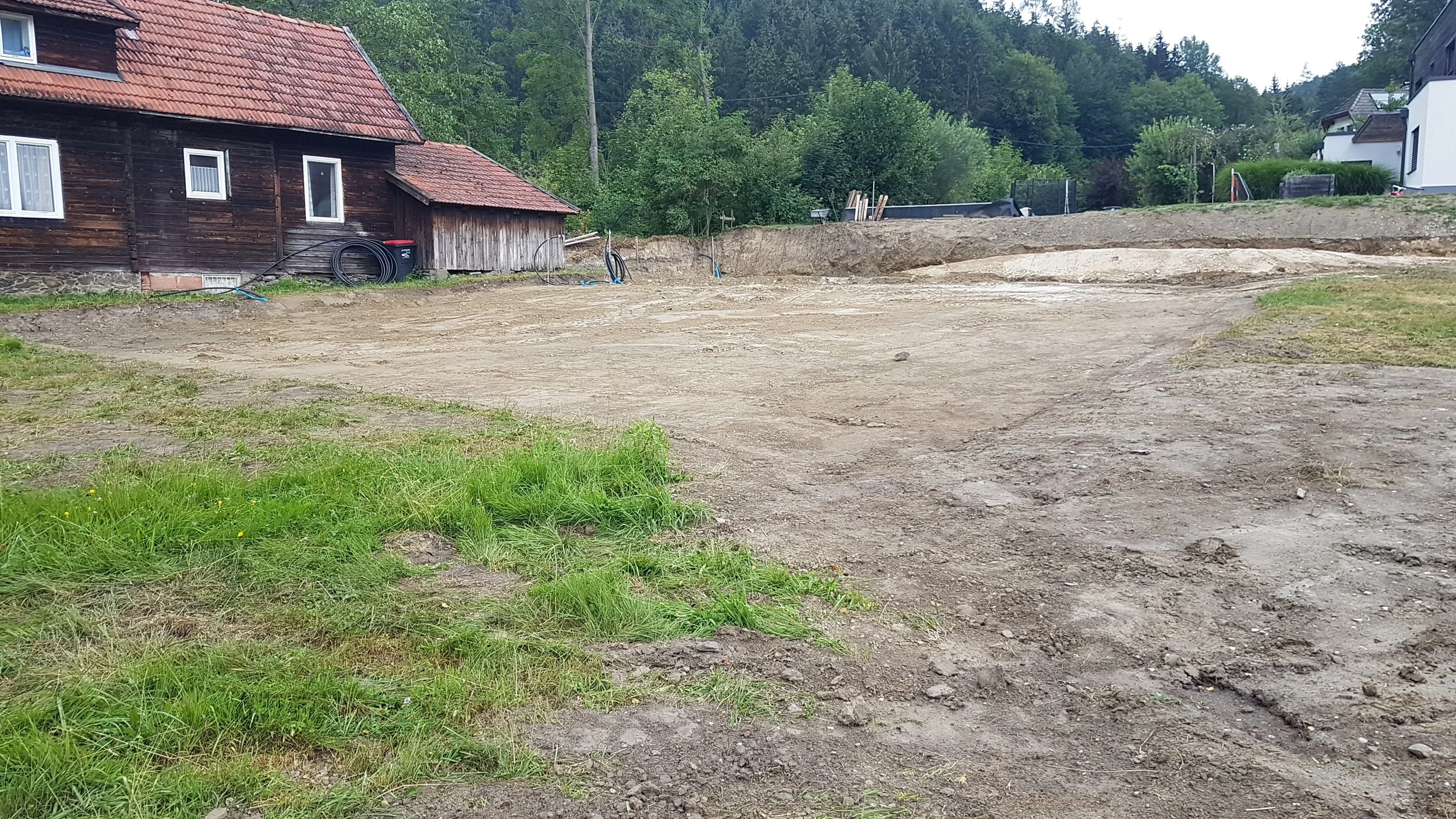 Baugrundstück mit aufbereitetem Boden, umgeben von Rasenflächen und einigen Häusern im Hintergrund, in einer ländlichen Umgebung mit Bäumen.