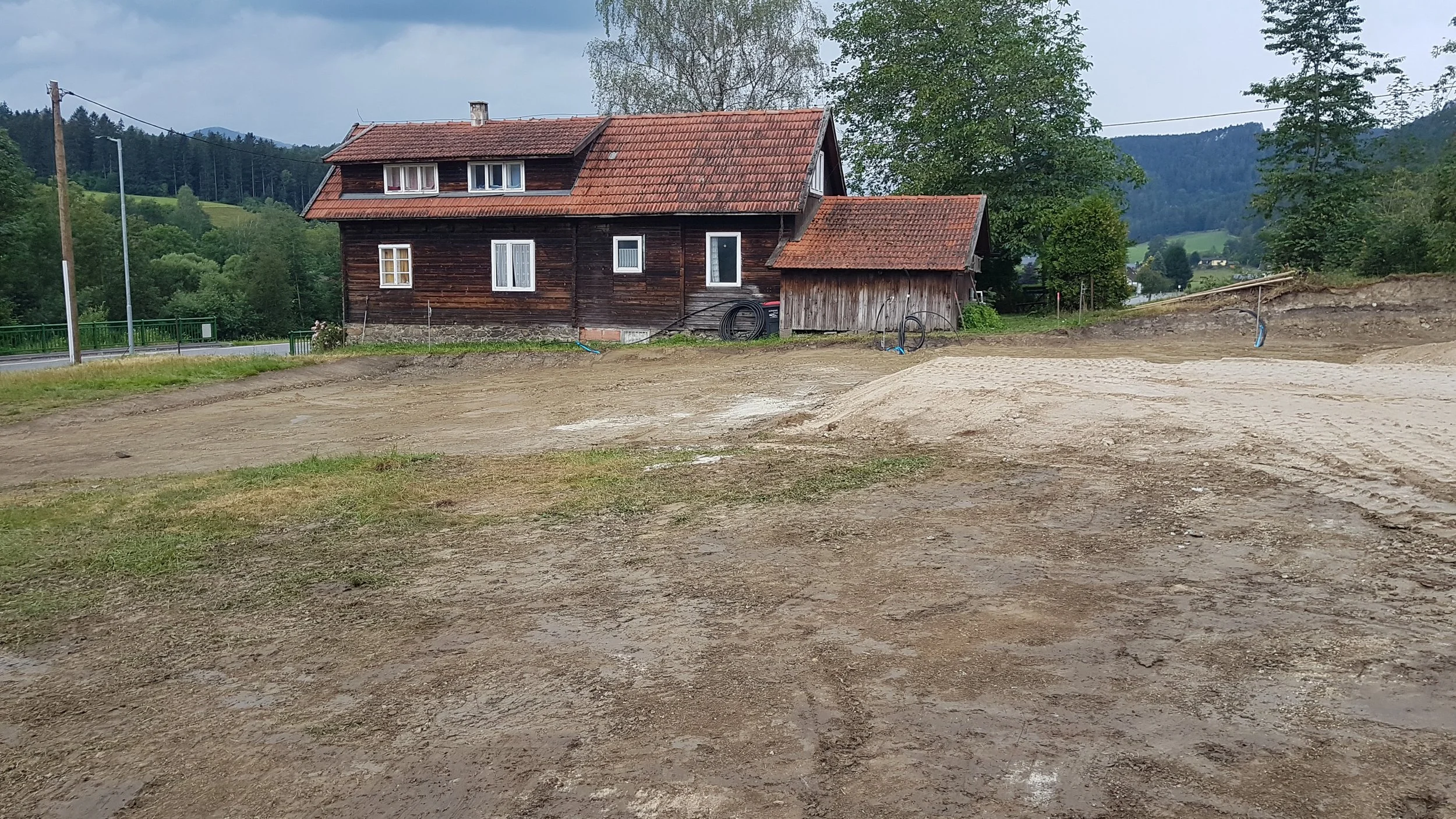 Unbebauter Boden vor einem alten, dunklen Holzhaus mit rotem Dach in einer ländlichen Umgebung mit Bäumen und Hügeln im Hintergrund.