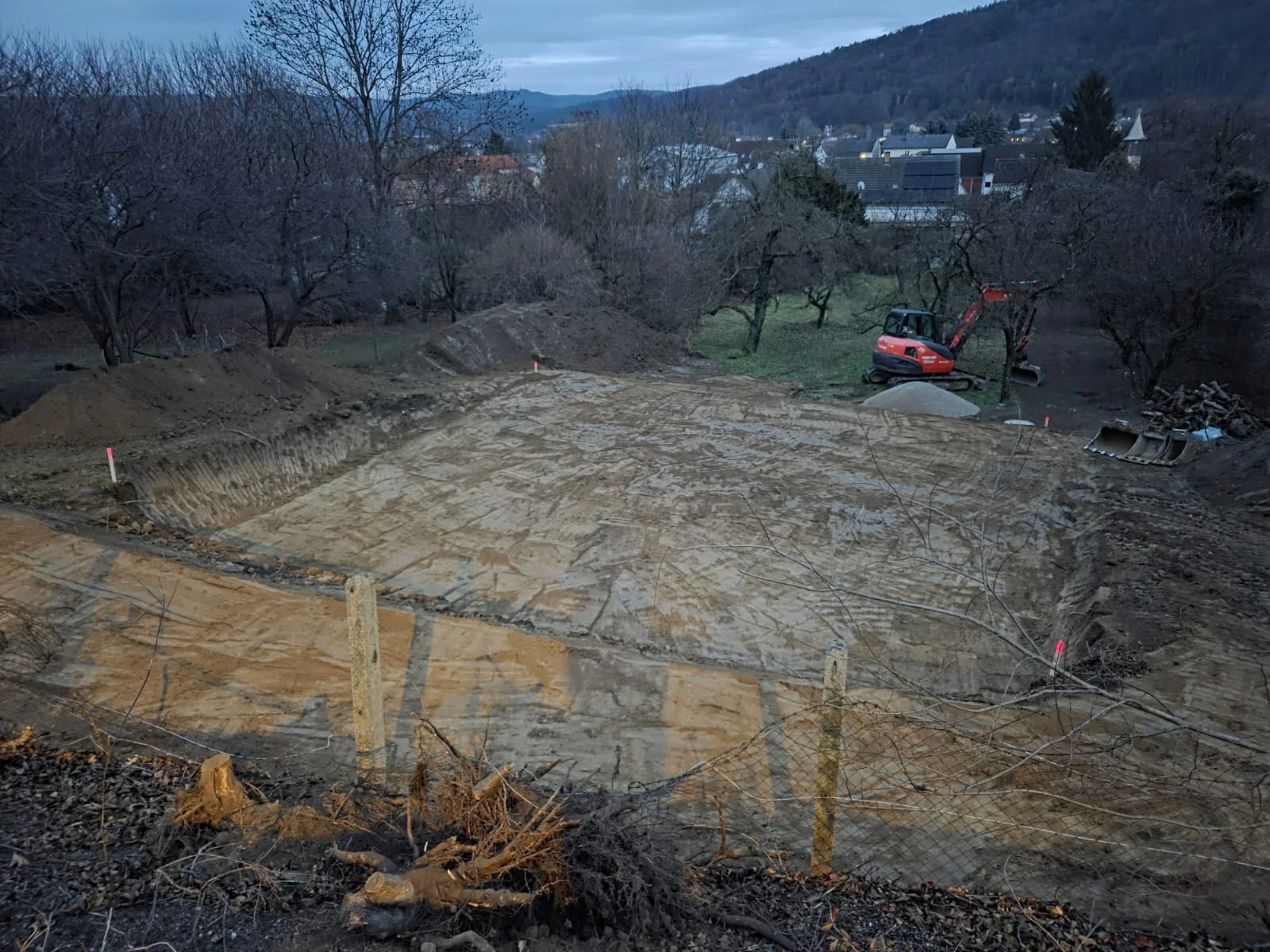 Baustelle mit Bagger, Erde und Markierungen, umgeben von Bäumen, in einem Tal mit Hügeln im Hintergrund.