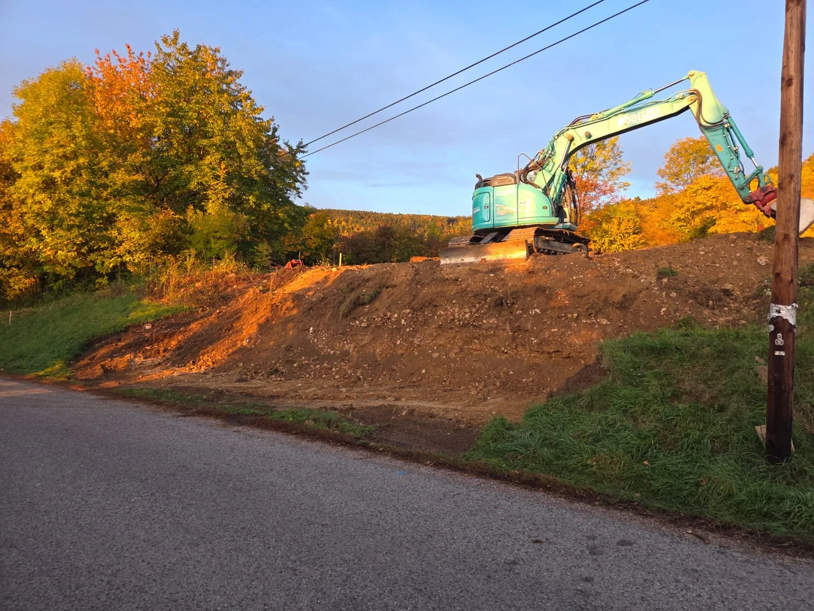 Ein Bagger arbeitet an einer Erschütterung neben einer Straße, umfahren von Bäumen bei Sonnenuntergang.