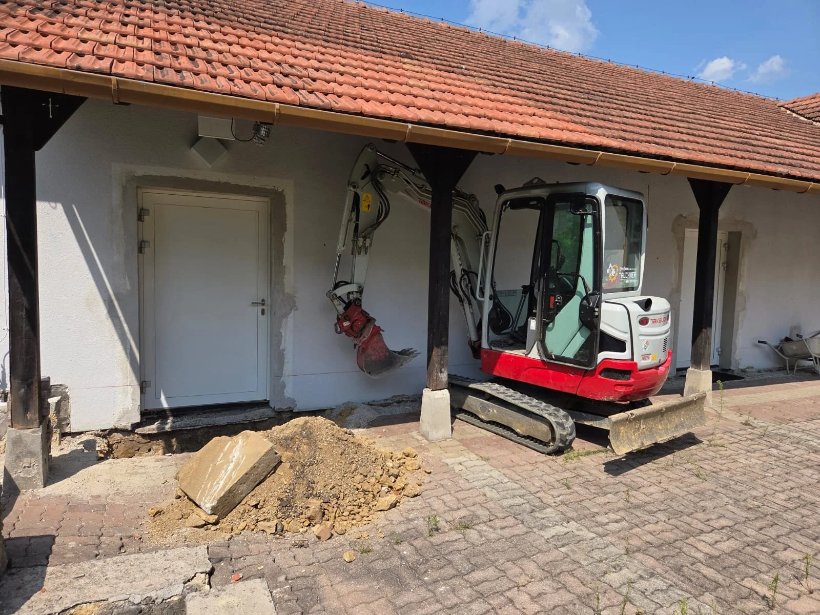 Kleiner Bagger im Einsatz vor einem weißen Gebäude mit rotem Ziegeldach, Baustelle mit Erde und Steinen im Vordergrund.