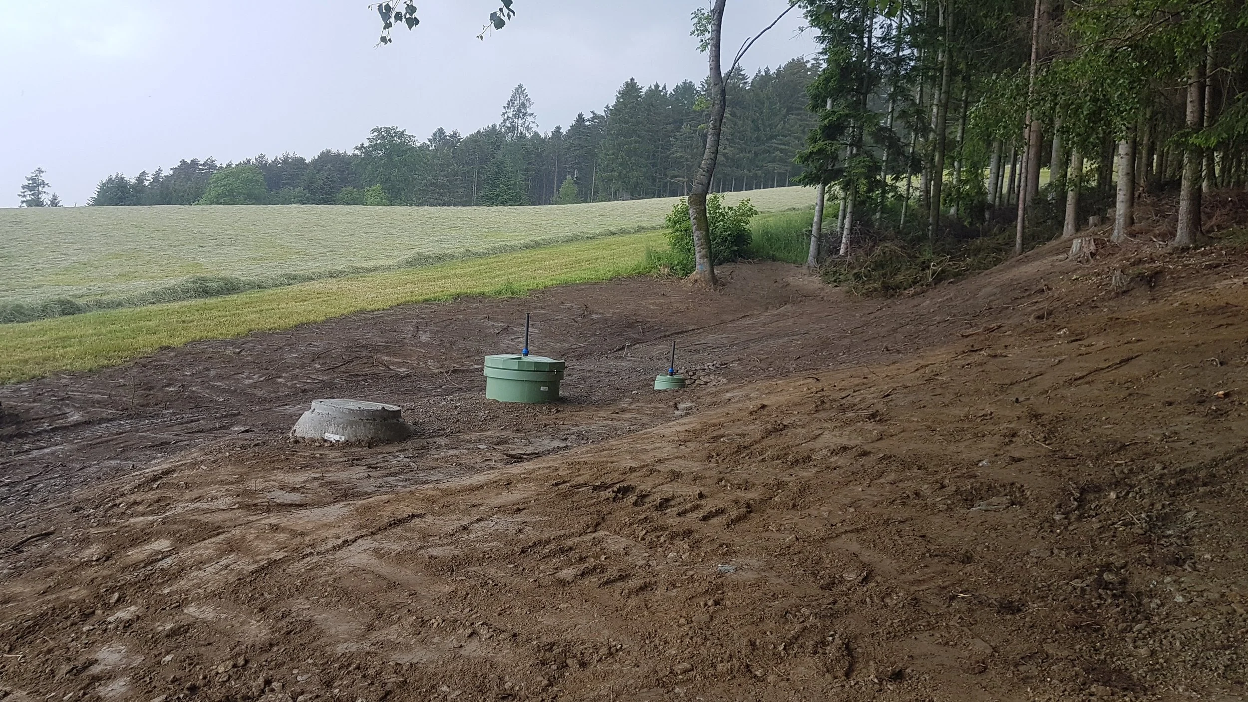 Boden mit offener Erde, im Hintergrund Wiese und Wald, zwei Gräben mit grünen Kontrollkästen und Wasserleitungen.